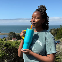 Load image into Gallery viewer, A woman stands with a reusable teal blue tumbler with The Marine Mammal Center logo and a scenic ocean background. She is acting like she is taking a sip from the tumbler.