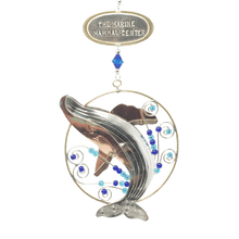 Load image into Gallery viewer, Silver and tan-colored metal ornament in shape of breaching humpback whale in a thin circular frame, with blue beads. Oval-shaped metal tag on top reads "THE MARINE MAMMAL CENTER".