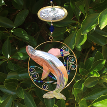 Load image into Gallery viewer, Silver and tan-colored metal ornament in shape of breaching humpback whale in a thin circular frame, with blue beads. Backdrop of green leaves.