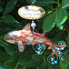 Load image into Gallery viewer, Copper and silver-colored metal orca and baby ornament with blue and white beads. Oval metal hangtag on hanger reads "THE MARINE MAMMAL CENTER". Backdrop of green leaves.