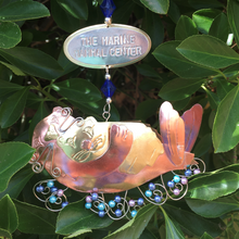 Load image into Gallery viewer, Copper and gold-colored metal ornament in shape of sea otter holding baby otter, with blue and purple beads. Oval-shaped metal tag on hanger reads "THE MARINE MAMMAL CENTER". Backdrop of green leaves.
