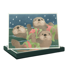 Load image into Gallery viewer, Box of holiday cards featuring three sea otters floating on ocean surface, each holding a red sea star and fronds of green kelp.