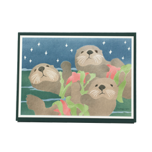 Load image into Gallery viewer, Box of holiday cards featuring three sea otters floating on ocean surface, each holding a red sea star and fronds of green kelp.