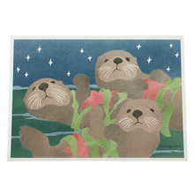 Load image into Gallery viewer, Holiday card featuring three sea otters floating on ocean surface, each holding a red sea star and fronds of green kelp.