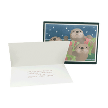 Load image into Gallery viewer, Open holiday card with inside message "May you have a Happy Holiday and a Joyous New Year". Backdrop is a box of holiday cards featuring three sea otters floating on ocean surface, each holding a red sea star and fronds of green kelp.