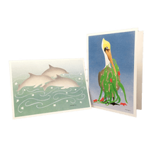 Load image into Gallery viewer, 2 holiday cards: 3 dolphins swimming against green-blue background dotted with white stars, and pelican holding fronds of kelp and red stars in its beak.