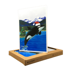 Load image into Gallery viewer, Holiday card with orca wearing Santa hat jumping out of water in front of a backdrop of mountains and a Christmas tree.