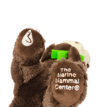Load image into Gallery viewer, Plush brown and light tan sea otter with embroidered eyes holding bright green kelp. Embroidered feet show white The Marine Mammal Center name and logo in white thread - view from the bottom of the plush.