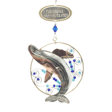 Load image into Gallery viewer, Silver and tan-colored metal ornament in shape of breaching humpback whale in a thin circular frame, with blue beads. Oval-shaped metal tag on top reads &quot;THE MARINE MAMMAL CENTER&quot;.
