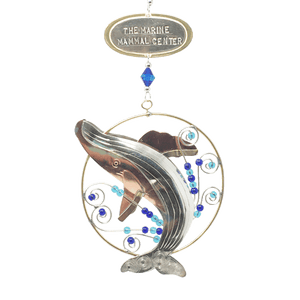 Silver and tan-colored metal ornament in shape of breaching humpback whale in a thin circular frame, with blue beads. Oval-shaped metal tag on top reads "THE MARINE MAMMAL CENTER".