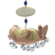 Copper and gold-colored metal ornament in shape of sea otter holding baby otter, with blue and purple beads. Oval-shaped metal tag on hanger reads 