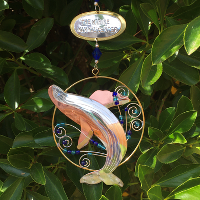 Silver and tan-colored metal ornament in shape of breaching humpback whale in a thin circular frame, with blue beads. Backdrop of green leaves. 