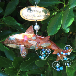 Copper and silver-colored metal orca and baby ornament with blue and white beads. Oval metal hangtag on hanger reads "THE MARINE MAMMAL CENTER". Backdrop of green leaves. 