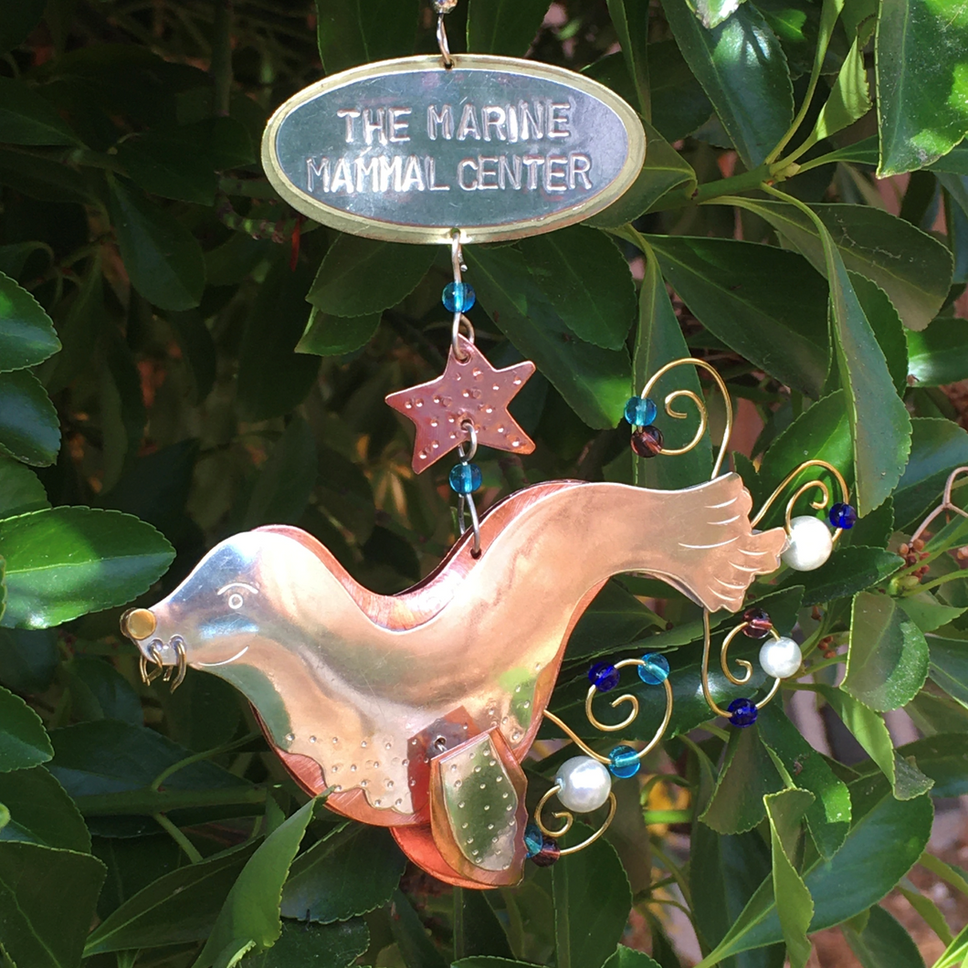 Copper and silver-colored metal ornament in shape of sea lion, with green, blue, and purple beads. Oval-shaped metal tang on hanger reads 