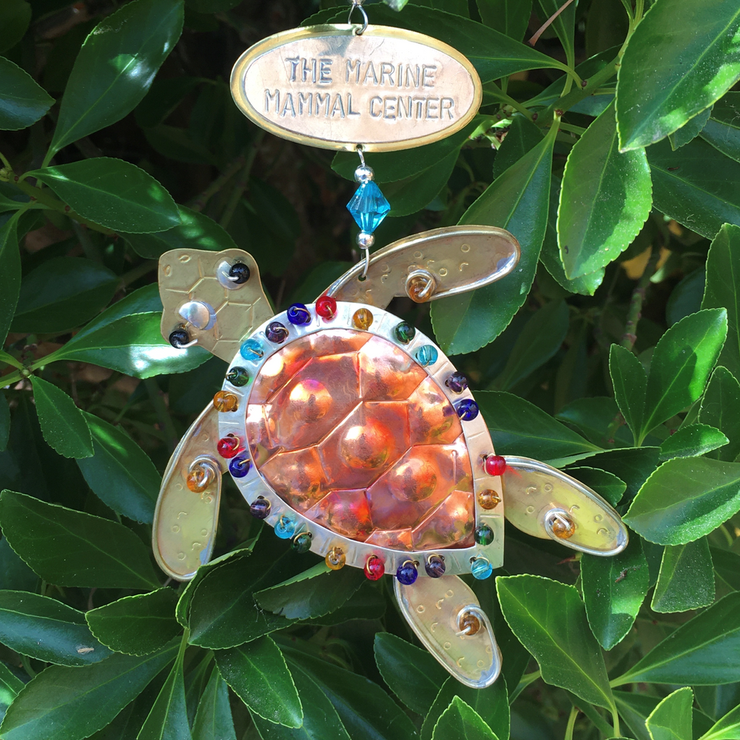 Copper and silver-colored metal sea turtle ornament with rainbow beads along edge of shell. Oval metal hangtag on hanger reads 