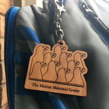 Load image into Gallery viewer, Wood keychain with laser-cut design of ten sea lion profiles and text &quot;The Marine Mammal Center&quot; underneath. Keychain hangs from a silver clip on a gray and blue backpack. 
