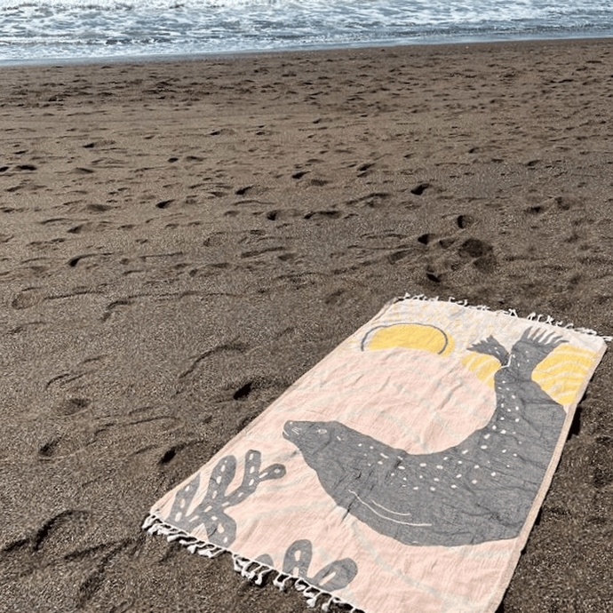 Peach and tan beach towel with harbor seal silhouette design, laid out on sand with ocean in background