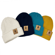 Load image into Gallery viewer, Four beanies lie splayed and overlapping in various colors, cream, navy, turquoise, and marigold, with cork tags on the cuff depicting The Marine Mammal Center Logo.
