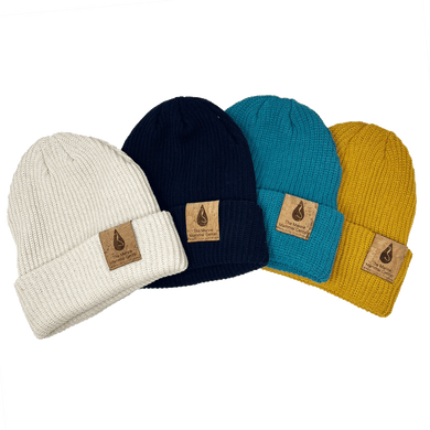 Four beanies lie splayed and overlapping in various colors, cream, navy, turquoise, and marigold, with cork tags on the cuff depicting The Marine Mammal Center Logo.