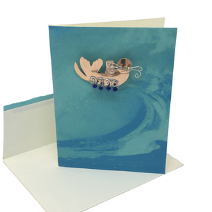 Blank greeting card with metal sea otter pin attached to light blue front cover, envelope below.