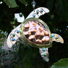 Load image into Gallery viewer, Silver and copper-colored metal ornament in shape of sea turtle, with blue, green, and white beads. Dark green leafy background.

