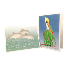 Load image into Gallery viewer, 2 holiday cards: 3 dolphins swimming against green-blue background dotted with white stars, and pelican holding fronds of kelp and red stars in its beak.
