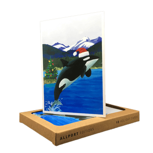Holiday card with orca wearing Santa hat jumping out of water in front of a backdrop of mountains and a Christmas tree.
