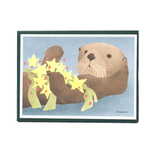 Load image into Gallery viewer, Holiday card box cover with sea otter holding yellow and red star ornaments and kelp.
