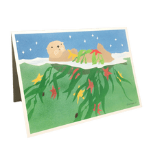 Load image into Gallery viewer, Holiday card with sea otter floating on green water under starry sky, holding fronds of kelp and red and yellow stars.
