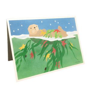 Holiday card with sea otter floating on green water under starry sky, holding fronds of kelp and red and yellow stars.