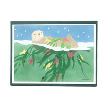 Load image into Gallery viewer, Holiday card box cover with sea otter floating in green water, holding kelp and red and yellow stars.
