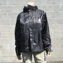 Load image into Gallery viewer, A headless mannequin stands in front of a grey brick wall. The mannequin is wearing a black, shiny jacket with a white TMMC upper left-hand chest logo and high collar.  The mannequin is also wearing light-wash jeans.
