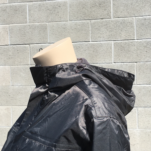 A headless mannequin's torso in profile stands in front of a grey brick wall, with only the bust and upward visible. The mannequin is wearing a black, shiny jacket with a hood and collar.   