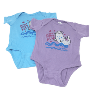 Purple and Blue "Sealed with a Kiss" onesies with The Marine Mammal Center name, depicting a cartoon seal grey with blue spots, small magenta hearts around the design, and blue wavey lines under the seal. The purple onesie is laying out flat slightly overlapping the blue onesie also laying out flat.