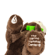 Load image into Gallery viewer, Plush brown and light tan sea otter with embroidered eyes holding bright green kelp. Embroidered feet show white The Marine Mammal Center name and logo in white thread - view from the bottom of the plush.
