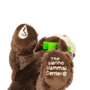 Plush brown and light tan sea otter with embroidered eyes holding bright green kelp. Embroidered feet show white The Marine Mammal Center name and logo in white thread - view from the bottom of the plush.