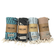 Load image into Gallery viewer, 4 Rolled beach towels with tassels of various colors, the towels are various colors blue, black, light blue, and sandy brown, wrapped in brown cardstock packaging reading &quot;Sand Cloud&quot;.
