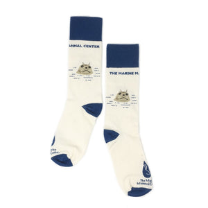 Two TMMC harbor seal logo socks, a mostly cream white sock with deep blue accents depicting TMMC logoing and harbor seal face popping out of water.