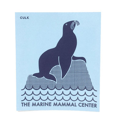 Light blue rectangular sticker with dark blue simplified design of a sea lion sitting on a rock in the waves of the ocean with 