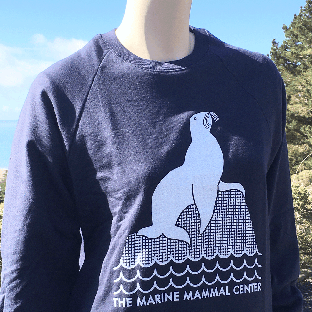 Navy blue sweatshirt with design in white of a sea lion perched on a rock above waves. Below the waves are the words THE MARINE MAMMAL CENTER in all caps. 