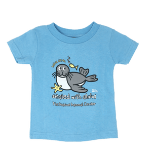 Light blue short sleeve t-shirt with a cartoon image of a grey and white monk seal with small yellow fish and seastar, words "Monk Seal" and "Sealed with Aloha" with the Center name and artist signature small in the corner.